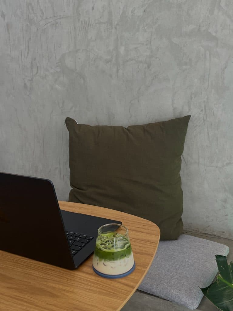 computer sitting on a desk next to a glass cup with matcha and a pillow sitting on a chair