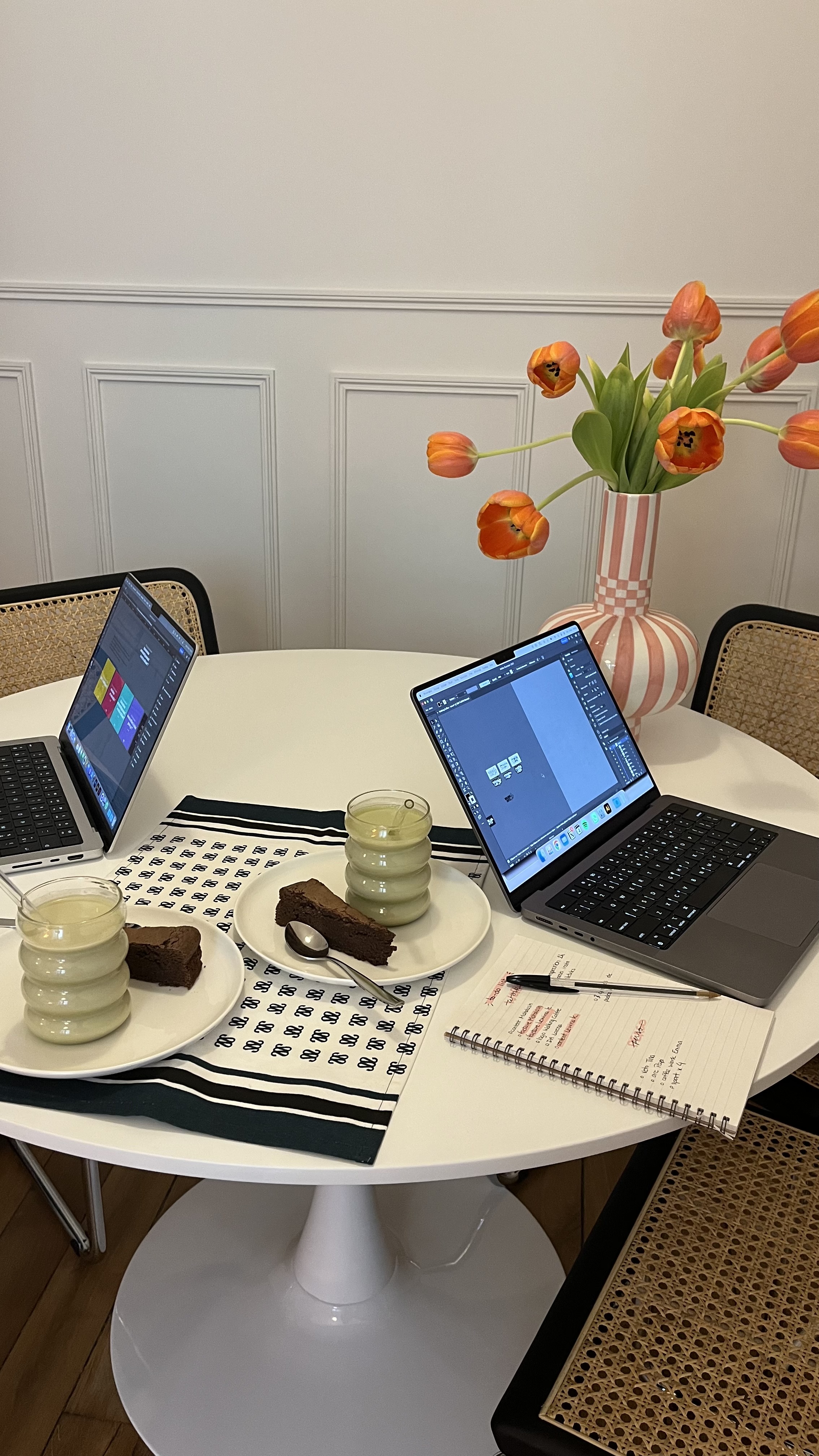 two computers sitting on a white desk with two glasses of matcha and chocolate cake and a floral arrangement in a vase