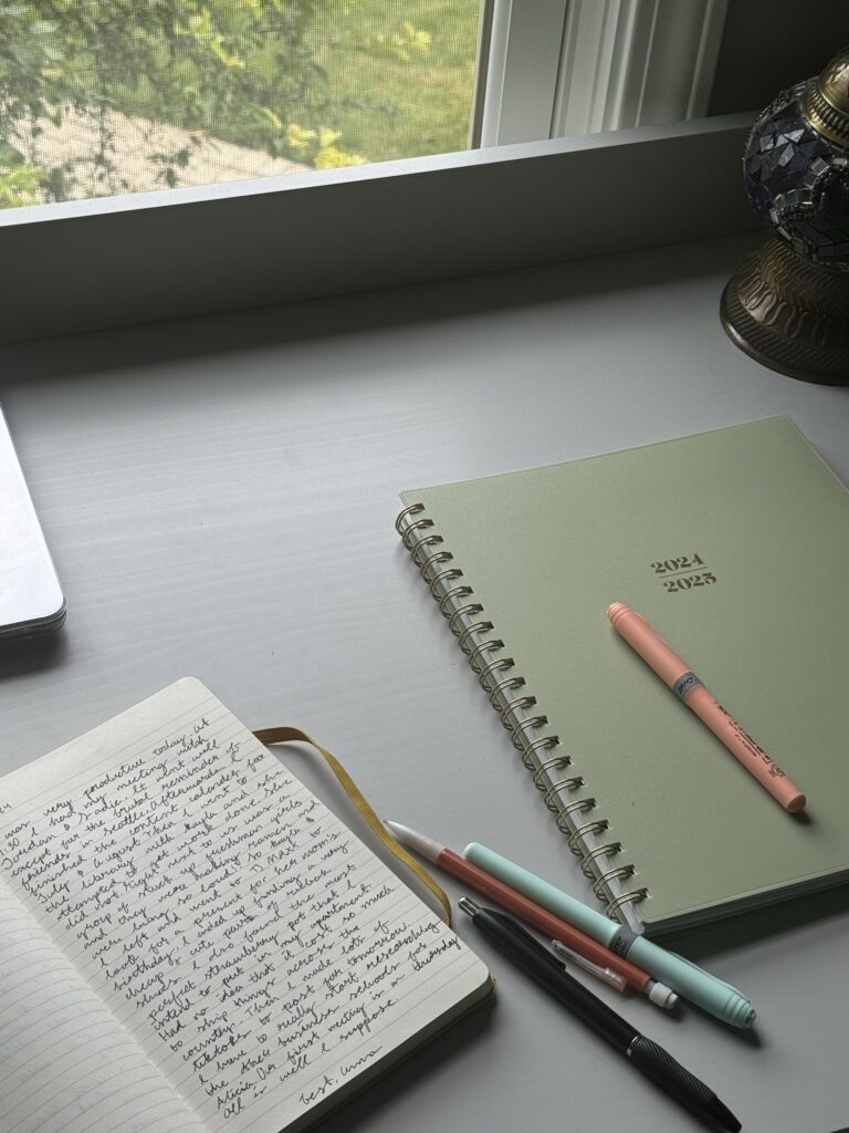 desk in an SEO company for small business office with open notebook of handwritten notes, green 2024 planner, scattered pens, laptop, and window plants