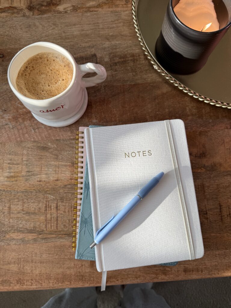 stack of notebooks on rustic wood desk beside coffee cup, blue pen, and burning candle