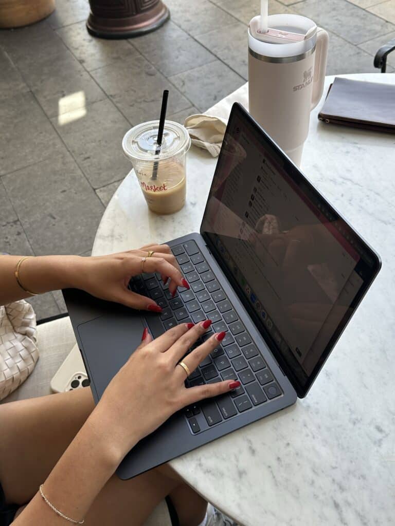 Woman with red nails types on MacBook at marble cafe table with iced Starbucks coffee and pink Stanley tumbler, evoking therapist branding