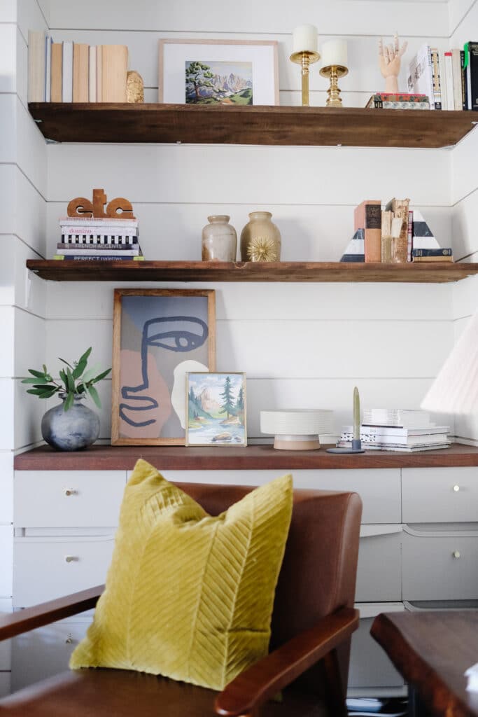 Cozy home workspace with wooden shelves, books, pottery, framed art, and a yellow cushion on a brown chair against a white paneled wall.