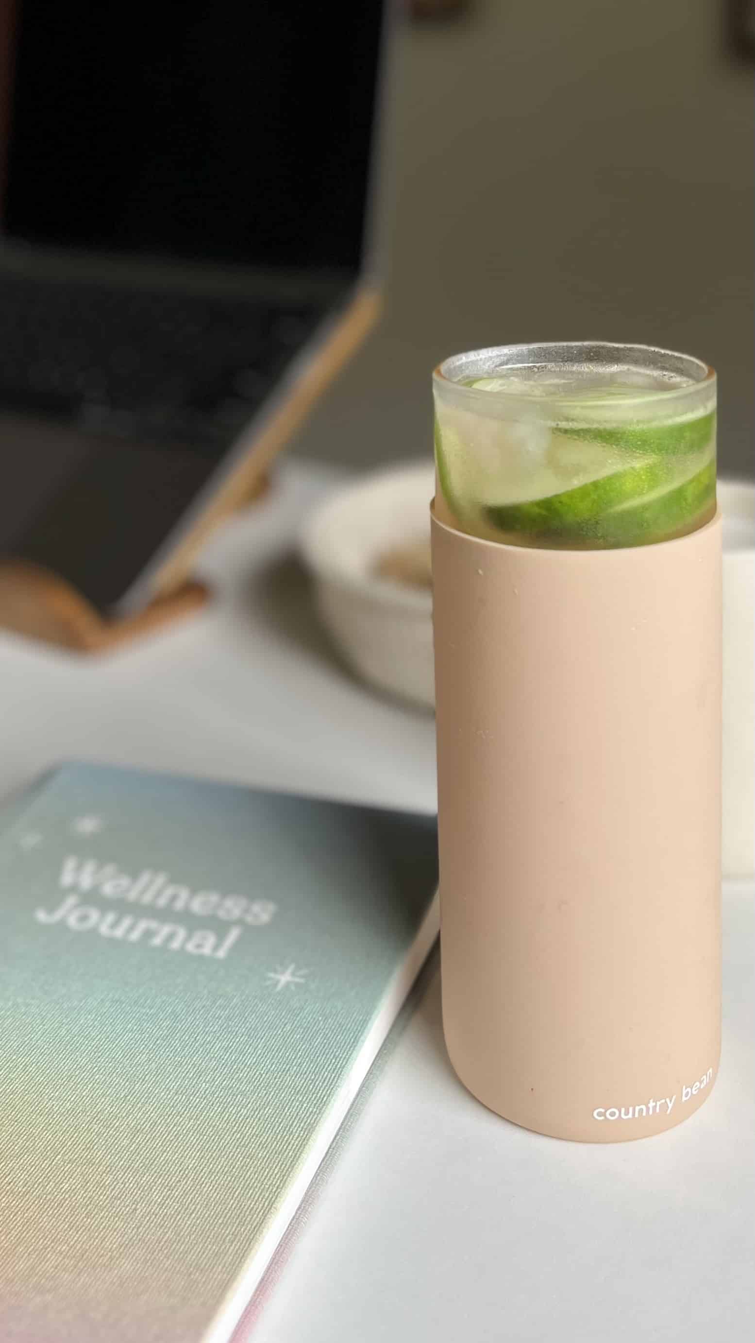 Blush-pink cylindrical bottle on a desk with a glass of lime-infused beverage resting on its rim.